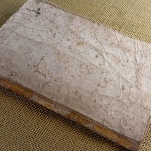 May include: A handmade, rectangular journal with a textured, off-white cover. The cover has a unique, organic appearance with visible fibres and imperfections. The spine is bound with a tan material. The journal is lying on a textured, tan surface.