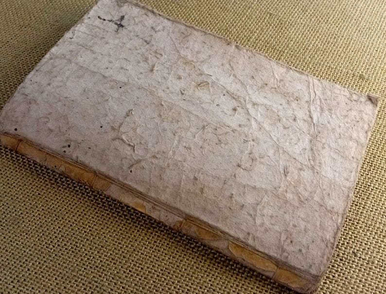 May include: A handmade, rectangular journal with a textured, off-white cover. The cover has a unique, organic appearance with visible fibres and imperfections. The spine is bound with a tan material. The journal is lying on a textured, tan surface.