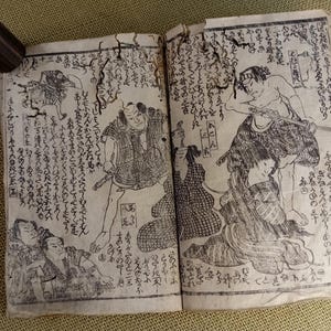 May include: An open antique book with Japanese illustrations and calligraphy. The black and white drawings depict figures in traditional clothing, with swords. A dark wooden cylinder rests on the book's edge.