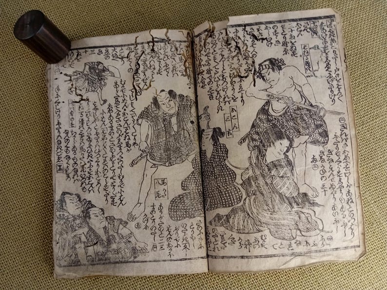 May include: An open antique book with Japanese illustrations and calligraphy. The black and white drawings depict figures in traditional clothing, with swords. A dark wooden cylinder rests on the book's edge.