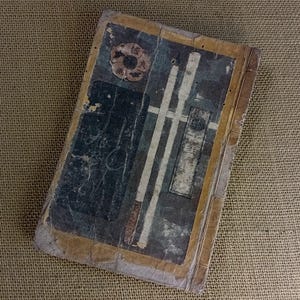 May include: An antique book with a distressed appearance. The cover displays faded blue, white, and brown illustrations, including a circular emblem and vertical stripes. The edges are yellowed, indicating age and use.