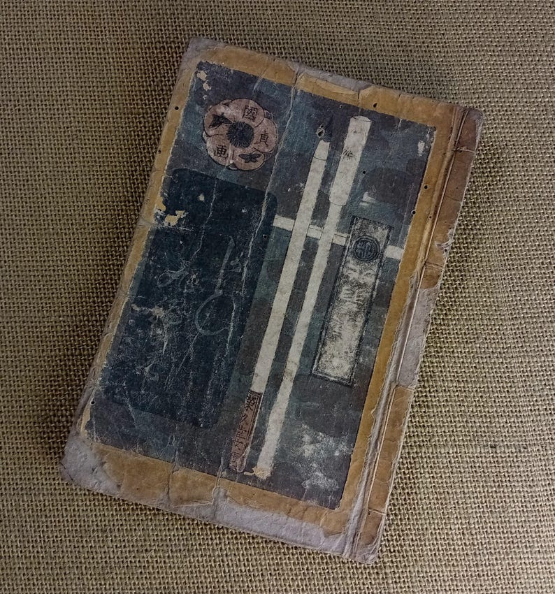 May include: An antique book with a distressed appearance. The cover displays faded blue, white, and brown illustrations, including a circular emblem and vertical stripes. The edges are yellowed, indicating age and use.