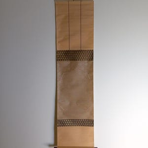 Op de afbeelding: Een rechthoekige, bruine rol met decoratieve donkerbruine randen en verticale lijnen. De rol hangt aan een witte muur. De onderkant van de rol is opgerold met rode accenten.