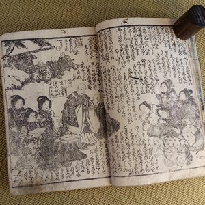 May include: An open antique book with detailed black and white illustrations and Japanese text. A dark wooden cylindrical object rests on the upper right page, possibly a bookmark or page weight. The book's pages show signs of age.