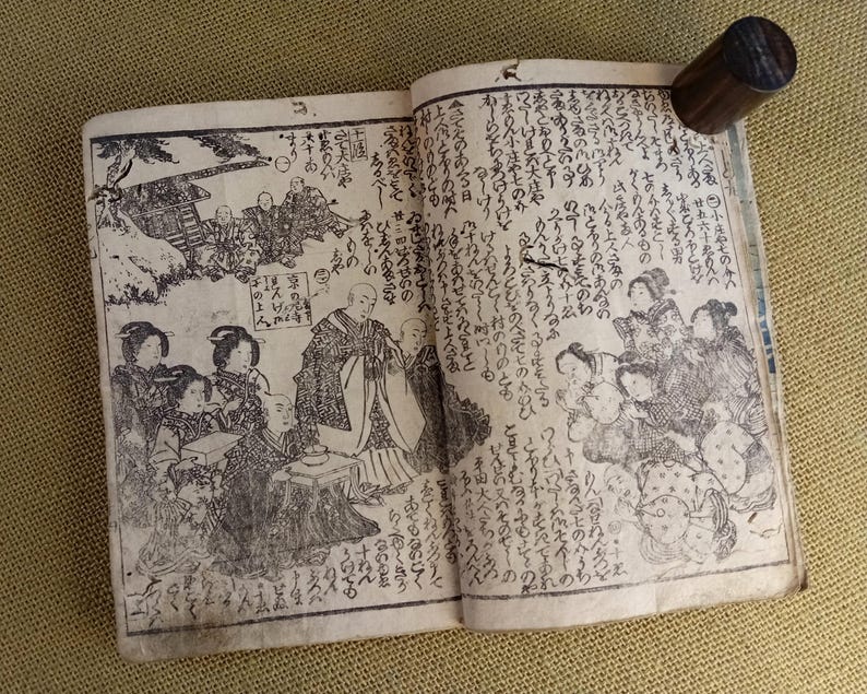 May include: An open antique book with detailed black and white illustrations and Japanese text. A dark wooden cylindrical object rests on the upper right page, possibly a bookmark or page weight. The book's pages show signs of age.