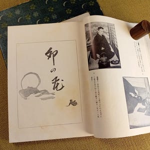 Op de afbeelding: Open boek met Japanse tekst en illustraties, waaronder een zwart-witfoto van een man. Een houten cilinder rust op de pagina. Het boek ligt op een getextureerd, goudbruin oppervlak.