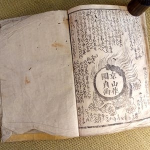 May include: An open antique book with Japanese text and illustrations. The right page features a circular emblem with Japanese characters and a turtle illustration. The pages are aged with visible wear and tear, suggesting its vintage.