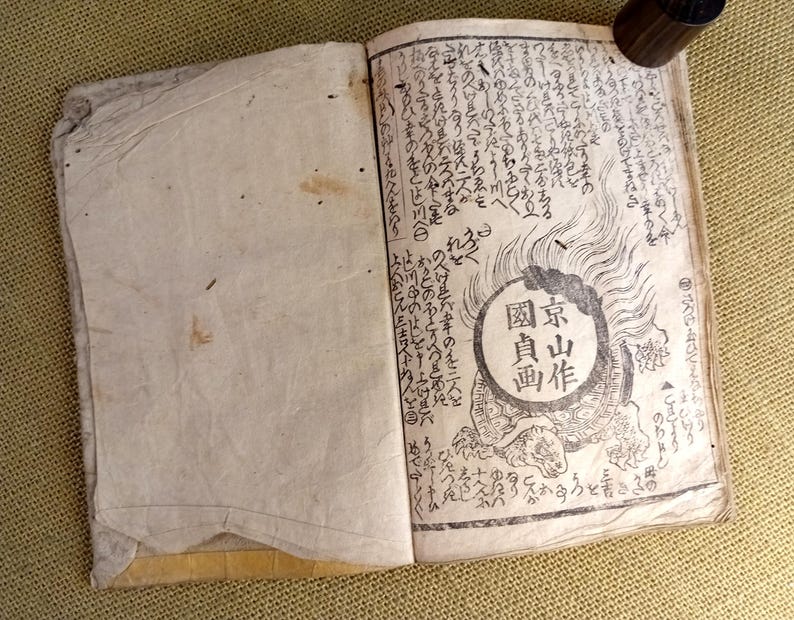 May include: An open antique book with Japanese text and illustrations. The right page features a circular emblem with Japanese characters and a turtle illustration. The pages are aged with visible wear and tear, suggesting its vintage.