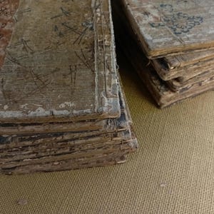 May include: Several stacks of aged, weathered books with worn, light-colored covers. The covers show faded red and black markings, along with handwritten notes and drawings. The pages are yellowed and appear fragile, suggesting historical significance.