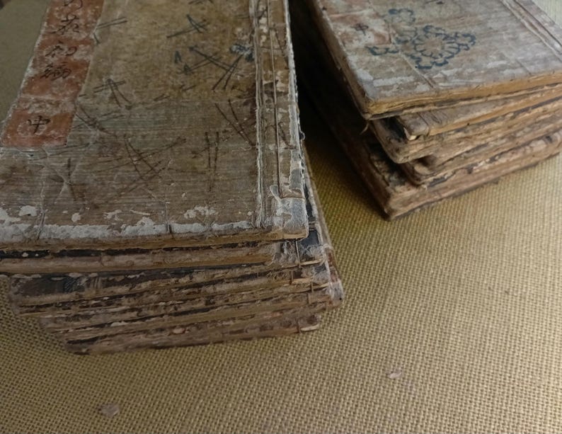 May include: Several stacks of aged, weathered books with worn, light-colored covers. The covers show faded red and black markings, along with handwritten notes and drawings. The pages are yellowed and appear fragile, suggesting historical significance.