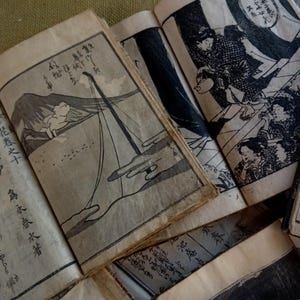 May include: A collection of antique Japanese woodblock print books, open to display detailed black and white illustrations and calligraphy. The pages are aged with visible wear, suggesting their historical significance. The illustrations depict scenes of people and landscapes.