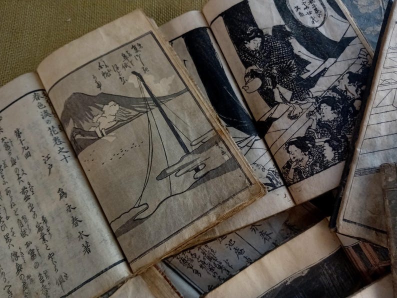 May include: A collection of antique Japanese woodblock print books, open to display detailed black and white illustrations and calligraphy. The pages are aged with visible wear, suggesting their historical significance. The illustrations depict scenes of people and landscapes.