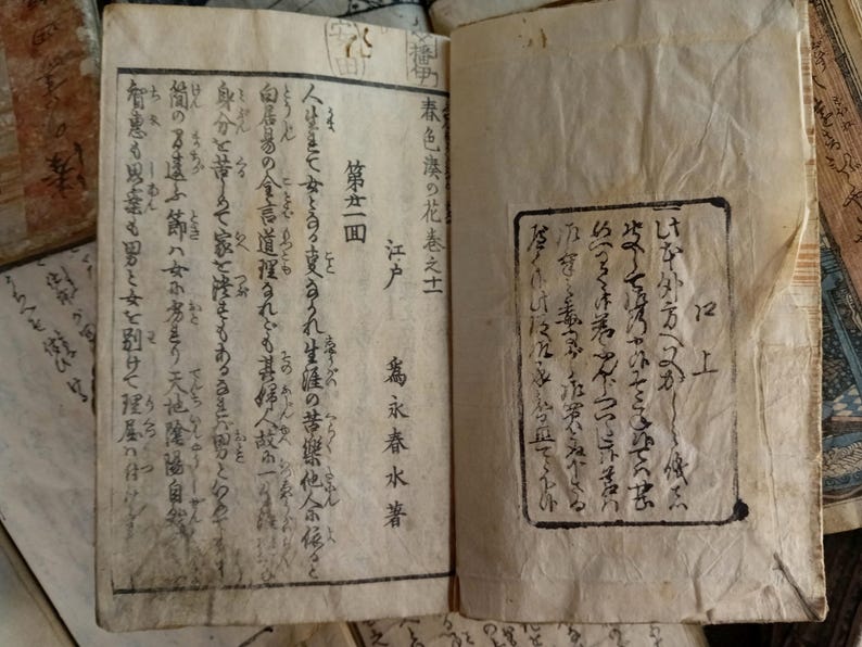 May include: An open book with aged, yellowed pages, featuring Japanese text and illustrations. The text is in black ink, with some areas appearing faded. The book is part of a collection of similar antique books.