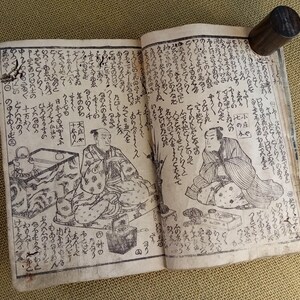May include: Open antique book with Japanese text and illustrations. The pages are aged and show detailed drawings of figures in traditional clothing. A dark wooden cylinder holds the pages open.