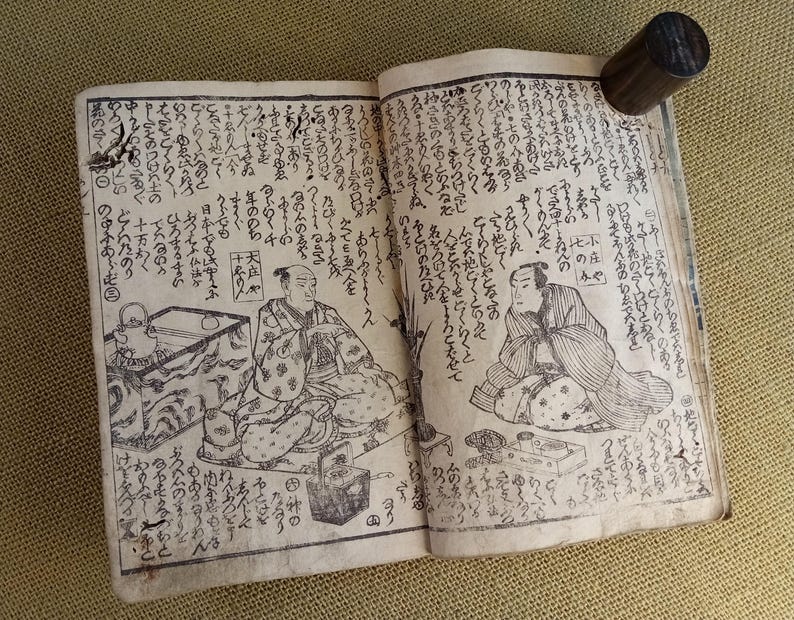May include: Open antique book with Japanese text and illustrations. The pages are aged and show detailed drawings of figures in traditional clothing. A dark wooden cylinder holds the pages open.