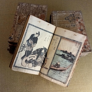 May include: Antique Japanese woodblock prints in a stack of books. The open book displays a scene with figures in traditional clothing and a river landscape. The covers are aged with visible wear and tear, showcasing the age of the books.