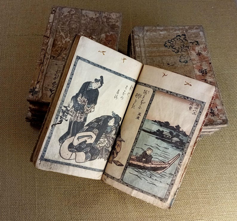 May include: Antique Japanese woodblock prints in a stack of books. The open book displays a scene with figures in traditional clothing and a river landscape. The covers are aged with visible wear and tear, showcasing the age of the books.