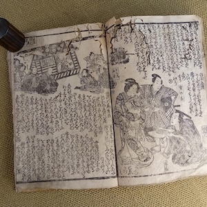 May include: An open antique book with Japanese text and illustrations. The pages are aged and worn, with detailed black and white drawings of people and scenes. A cylindrical, dark wooden object rests on the book's edge.