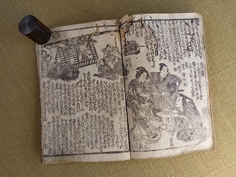 May include: An open antique book with Japanese text and illustrations. The pages are aged and worn, with detailed black and white drawings of people and scenes. A cylindrical, dark wooden object rests on the book's edge.