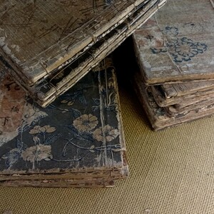 May include: A collection of antique books with aged, textured covers. The books are bound with wire and feature faded floral and geometric designs in muted colors. Some covers display Asian characters. The overall aesthetic is vintage and weathered.