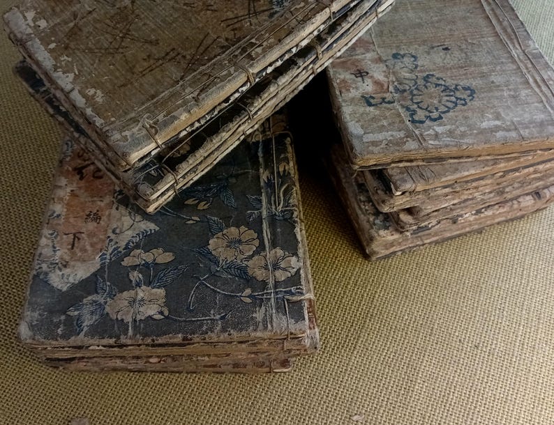 May include: A collection of antique books with aged, textured covers. The books are bound with wire and feature faded floral and geometric designs in muted colors. Some covers display Asian characters. The overall aesthetic is vintage and weathered.