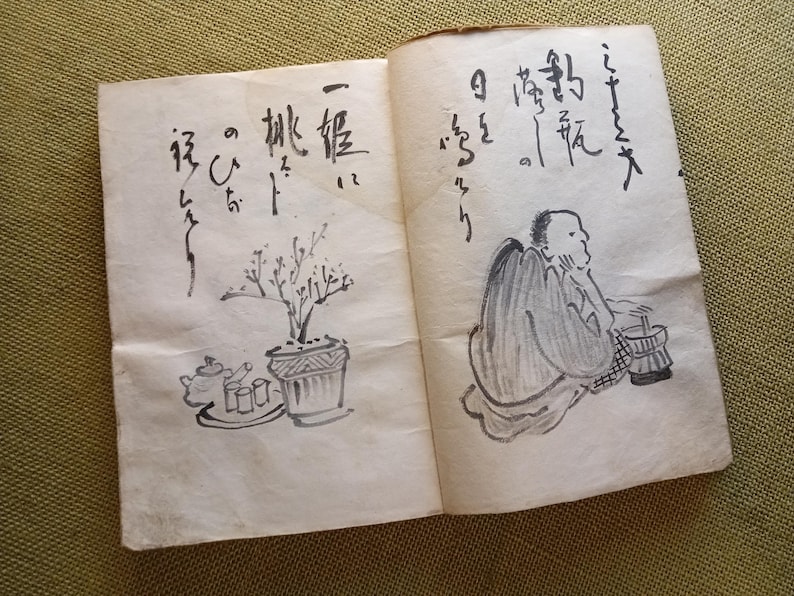 Puede incluir: Libro abierto con caligraf&iacute;a japonesa y dibujos a tinta. Una p&aacute;gina presenta una planta en maceta y un juego de t&eacute;. La otra p&aacute;gina muestra una figura sentada. Las p&aacute;ginas est&aacute;n envejecidas con un tono amarillento.