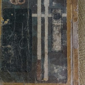 May include: Close-up of a distressed antique book cover. The cover has a dark background with vertical white stripes and Japanese text. A circular emblem and other Japanese characters are visible, with a worn appearance.