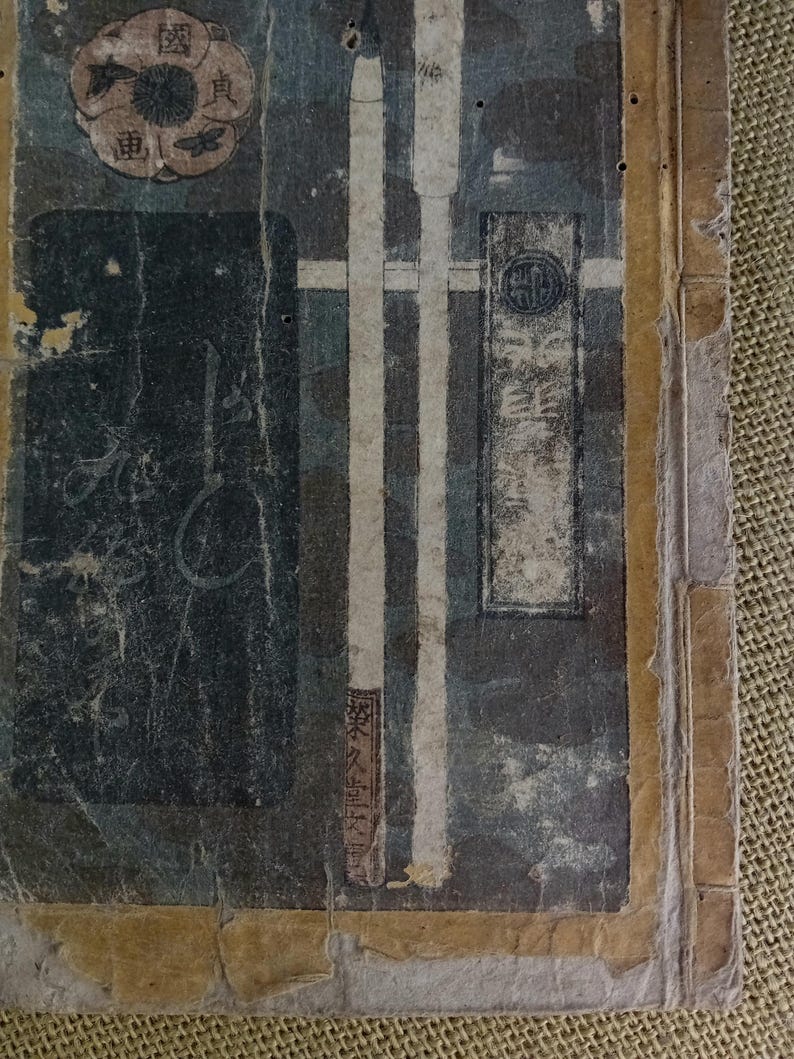 May include: Close-up of a distressed antique book cover. The cover has a dark background with vertical white stripes and Japanese text. A circular emblem and other Japanese characters are visible, with a worn appearance.