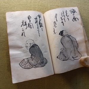 Puede incluir: Un libro antiguo abierto con caligraf&iacute;a japonesa y dibujos a tinta de figuras. Los dibujos son en tinta negra sobre papel envejecido blanquecino. El libro est&aacute; abierto sobre una superficie verde.