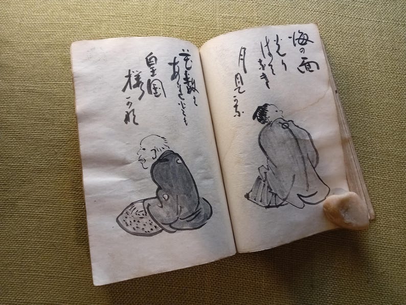 Puede incluir: Un libro antiguo abierto con caligraf&iacute;a japonesa y dibujos a tinta de figuras. Los dibujos son en tinta negra sobre papel envejecido blanquecino. El libro est&aacute; abierto sobre una superficie verde.