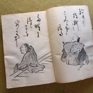 Puede incluir: Libro abierto con caligraf&iacute;a japonesa e ilustraciones a tinta de figuras. Los dibujos son en tinta negra sobre papel envejecido. La p&aacute;gina izquierda muestra una figura sentada de perfil. La p&aacute;gina derecha representa una figura de espaldas.