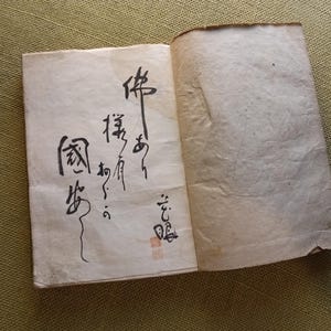 Puede incluir: Un libro antiguo abierto, con p&aacute;ginas envejecidas de color blanquecino. La p&aacute;gina izquierda presenta una elegante caligraf&iacute;a negra en escritura vertical, con un sello rojo en la parte inferior. La p&aacute;gina derecha est&aacute; en blanco. El libro es probablemente de origen asi&aacute;tico.