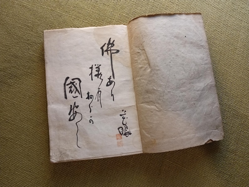 Puede incluir: Un libro antiguo abierto, con p&aacute;ginas envejecidas de color blanquecino. La p&aacute;gina izquierda presenta una elegante caligraf&iacute;a negra en escritura vertical, con un sello rojo en la parte inferior. La p&aacute;gina derecha est&aacute; en blanco. El libro es probablemente de origen asi&aacute;tico.