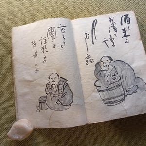 Puede incluir: Un libro abierto con caligraf&iacute;a e ilustraciones japonesas. Los dibujos representan figuras cerca de barriles y recipientes. Las p&aacute;ginas est&aacute;n envejecidas, con una peque&ntilde;a piedra clara en la esquina inferior izquierda.