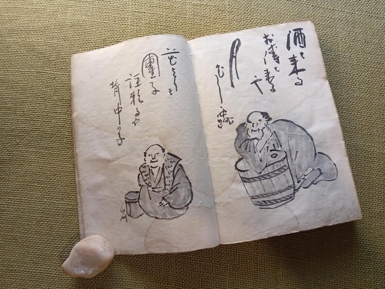 Puede incluir: Un libro abierto con caligraf&iacute;a e ilustraciones japonesas. Los dibujos representan figuras cerca de barriles y recipientes. Las p&aacute;ginas est&aacute;n envejecidas, con una peque&ntilde;a piedra clara en la esquina inferior izquierda.