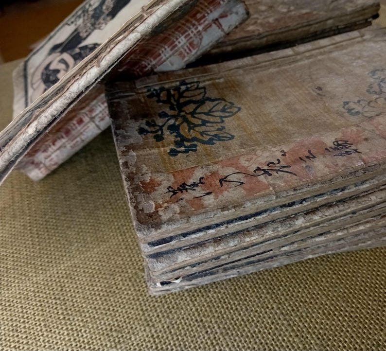 May include: A stack of aged, rectangular paper sheets with faded colors and visible wear. The top sheet features a floral design in blue and black, with Japanese characters. The edges show signs of age and use, suggesting a vintage or antique item.