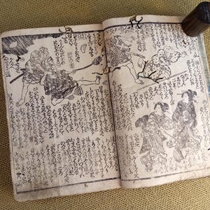 May include: An open antique book featuring Japanese illustrations and calligraphy. The pages are filled with detailed black and white drawings of figures in traditional attire, accompanied by handwritten text. A dark wooden cylinder rests on the top right page.