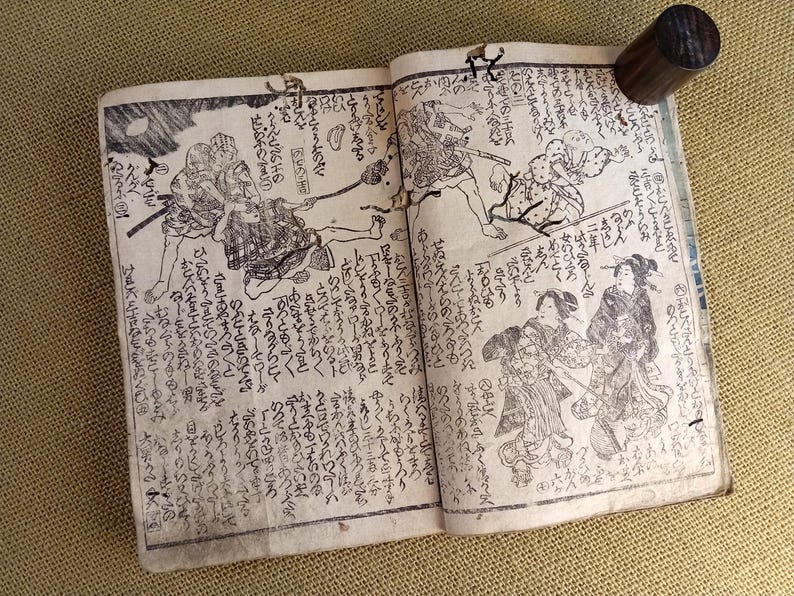 May include: An open antique book featuring Japanese illustrations and calligraphy. The pages are filled with detailed black and white drawings of figures in traditional attire, accompanied by handwritten text. A dark wooden cylinder rests on the top right page.