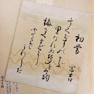 May include: A square piece of off-white paper featuring black Japanese calligraphy written in vertical columns. The paper has a subtle, wavy, light-gold decorative pattern across the centre and includes additional small text in the bottom corner.