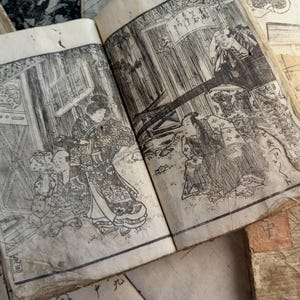 May include: Open book with detailed black and white illustrations of Japanese figures in traditional clothing. The pages are aged and worn, with visible signs of use. Other aged paper items are in the background.