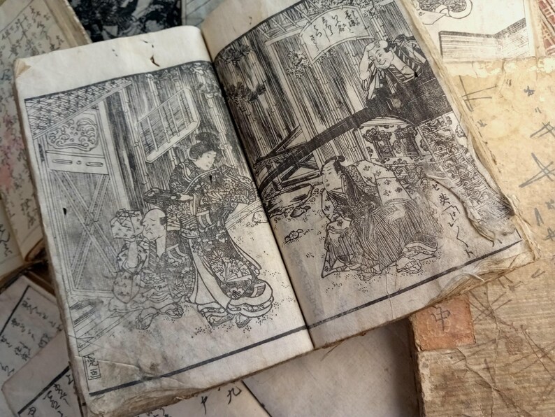 May include: Open book with detailed black and white illustrations of Japanese figures in traditional clothing. The pages are aged and worn, with visible signs of use. Other aged paper items are in the background.