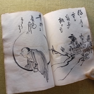 Puede incluir: Un libro abierto con pinturas tradicionales japonesas con tinta. La p&aacute;gina izquierda presenta una figura dentro de un c&iacute;rculo, mientras que la p&aacute;gina derecha representa un paisaje con edificios y &aacute;rboles. Caligraf&iacute;a presente.
