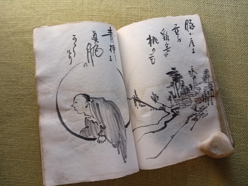 Puede incluir: Un libro abierto con pinturas tradicionales japonesas con tinta. La p&aacute;gina izquierda presenta una figura dentro de un c&iacute;rculo, mientras que la p&aacute;gina derecha representa un paisaje con edificios y &aacute;rboles. Caligraf&iacute;a presente.