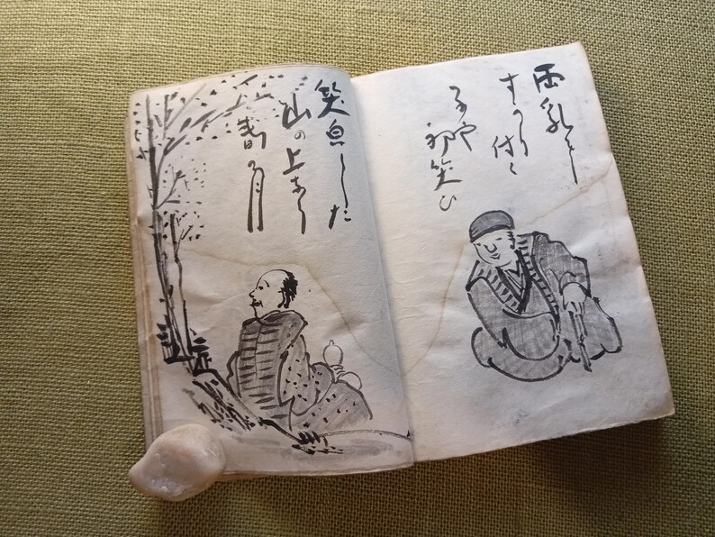 Puede incluir: Un libro abierto con caligraf&iacute;a japonesa e ilustraciones a tinta de figuras y &aacute;rboles. Las p&aacute;ginas est&aacute;n envejecidas, con un tono beige claro. Las ilustraciones son en tinta negra, representando escenas de personas y naturaleza.