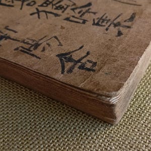 Puede incluir: Primer plano de un libro antiguo con una cubierta marr&oacute;n desgastada. Las p&aacute;ginas est&aacute;n envejecidas y la cubierta tiene caligraf&iacute;a negra. La esquina del libro es visible, mostrando el grosor de las p&aacute;ginas. El libro descansa sobre una superficie texturizada.