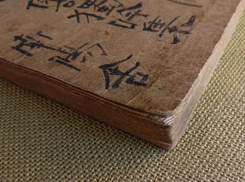 Puede incluir: Primer plano de un libro antiguo con una cubierta marr&oacute;n desgastada. Las p&aacute;ginas est&aacute;n envejecidas y la cubierta tiene caligraf&iacute;a negra. La esquina del libro es visible, mostrando el grosor de las p&aacute;ginas. El libro descansa sobre una superficie texturizada.