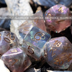 Op de afbeelding: Een set polyhedrale dobbelstenen met een doorschijnende, rokerige paarse tint en ingebed iriserende glitters. Elke dobbelsteen heeft gegraveerde goudkleurige cijfers. De tekst ALLUREDICE wordt herhaald in een horizontale band over het midden.