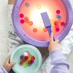 Könnte beinhalten: Ein Kind spielt mit einem leuchtenden lila Spielzeug, das eine durchsichtige Plastikschale mit bunten, durchsichtigen Zählern hat. Das Spielzeug wird mit einem magnetischen Zauberstab geliefert. Das Kind hält auch eine grüne Schale mit weiteren Zählern.