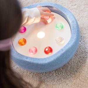 Könnte beinhalten: Ein leuchtendes sensorisches Spielzeug mit blauem Rand und weißer Mitte. In der Mitte befinden sich farbige, durchscheinende Edelsteine in Rosa-, Orange-, Grün- und Rottönen. Eine Kinderhand greift nach den Edelsteinen. Für sensorische Erkundung.