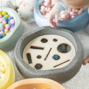 Könnte beinhalten: Eine Sammlung farbenfroher sensorischer Spielzeuge für Kleinkinder. Das Bild zeigt mehrere Schalen mit Pompons in verschiedenen Farben und eine graue Schale mit einem Holzeinsatz mit ausgeschnittenen Formen. Die Spielzeuge sind für die frühkindliche Entwicklung konzipiert.